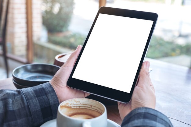 mani della donna che tengono tablet pc nero con schermo vuoto con tazza di caffè sulla tavola di legno nella caffetteria