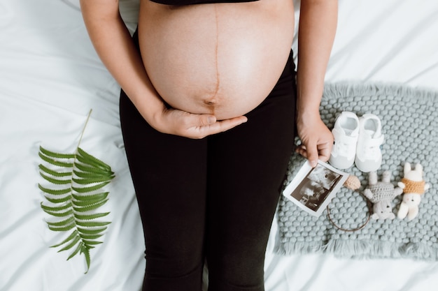 Mani della donna incinta che si tengono sulla pancia facendo un simbolo di cura e amore sulla camera da letto con immagine ad ultrasuoni. Madre in attesa in attesa e preparazione per la nascita del bambino. Concetto di donna incinta.