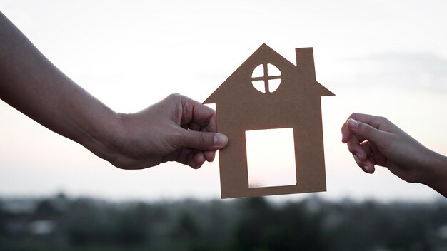 Mani della figlia e della madre del primo piano che tengono insieme la casa di carta casa contro il tramonto con il fuoco molle