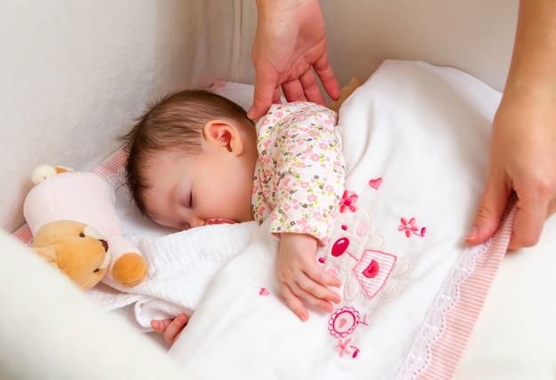 Mani della madre che accarezzano la sua graziosa bambina che dorme in un lettino con ciuccio e peluche