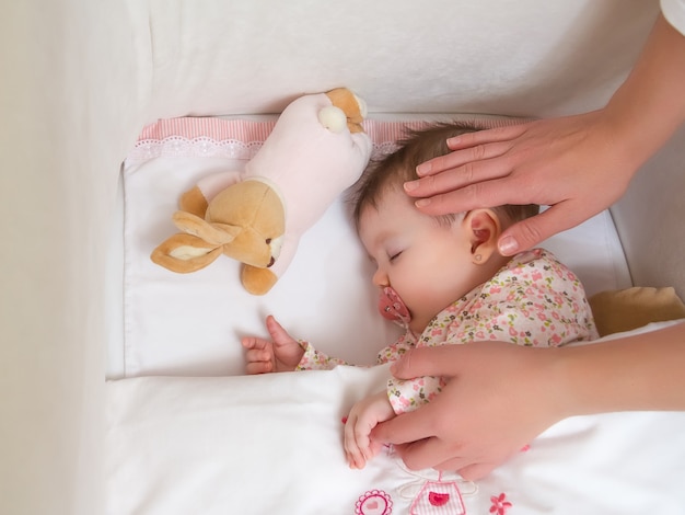 Mani della madre che accarezzano la sua graziosa bambina che dorme in un lettino con ciuccio e peluche