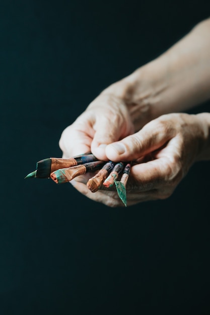 Mani della vecchia artista femminile che tengono pennelli sporchi e disordinati con diverse vernici in studio d'arte. Stile di vita e concetto di hobby