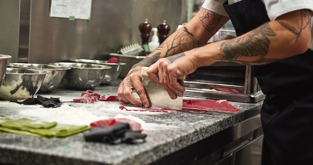 mani dello chef con tatuaggi che tagliano pasta sul tavolo con farina per pasta fatta in casa in cucina