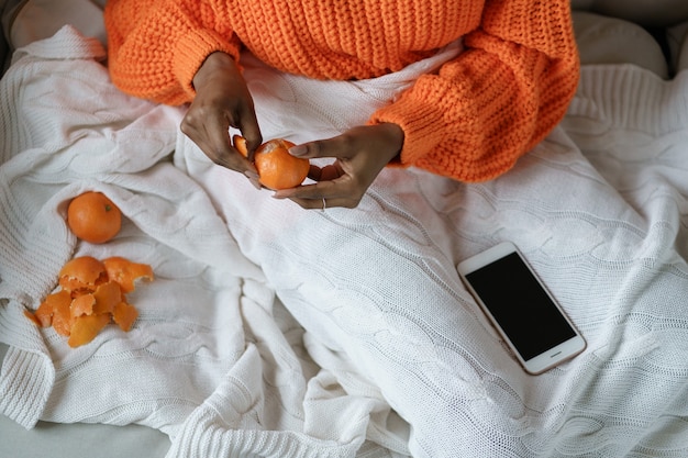 Mani di donna afro che sbucciano il mandarino dolce maturo, indossano un maglione arancione, sdraiato a letto sotto il plaid bianco lavorato a maglia. Frutta invernale, concetto di Natale.