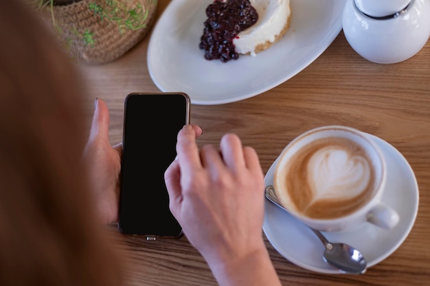 Mani di donna con smartphone in caffetteria contro tavolo cappuccino caffè e dessert Smartphone in mani femminili Lavoro durante il pranzo
