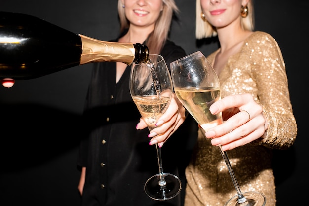 Mani di due giovani donne eleganti che tengono flauti per champagne mentre qualcuno versa la bevanda dalla bottiglia alla festa in discoteca