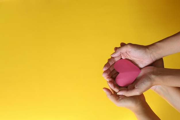 Mani di due persone che tengono una forma di cuore rosso su sfondo giallo con spazio di copia.