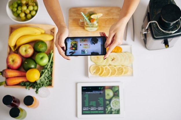 Mani di food blogger phototgraphing tavolo da cucina con piatto di frutta a fette, tavoletta digitale con ricetta frullato verde e frullatore