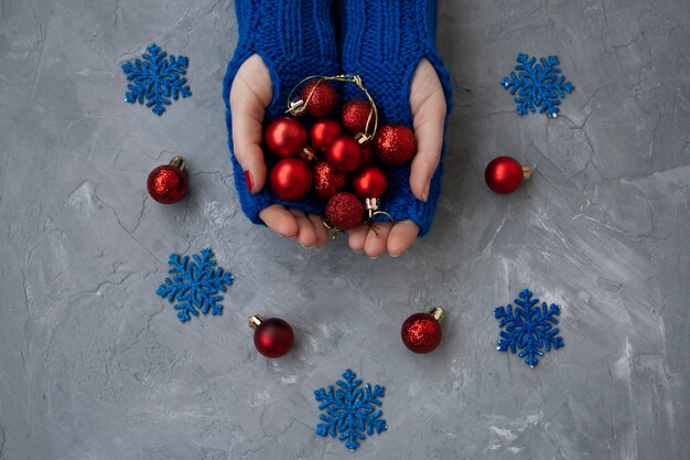 Mani di giovane bella donna in guanti blu. Tiene le palle rosse di Natale. Nelle vicinanze si trovano fiocchi di neve blu e decorazioni rosse sull'albero di Natale.