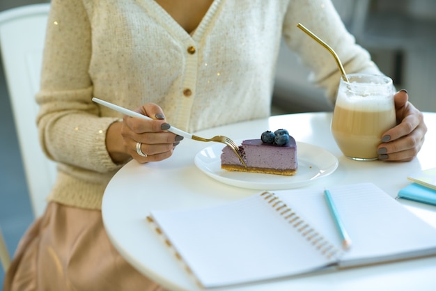 Mani di giovane donna casual prendendo un pezzo di gustosa cheesecake ai mirtilli e avendo cappuccino seduti nella caffetteria