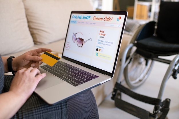 Mani di giovane donna disabile contemporanea che tiene la carta di credito sulla tastiera del laptop mentre va a fare ordine nel negozio online da casa