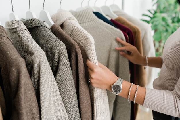 Mani di giovane donna shopper in pullover grigio guardando attraverso la nuova collezione stagionale di abbigliamento casual in boutique