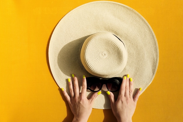 Mani di giovane ragazza che detiene cappello di paglia e occhiali da sole su sfondo giallo vibrante.