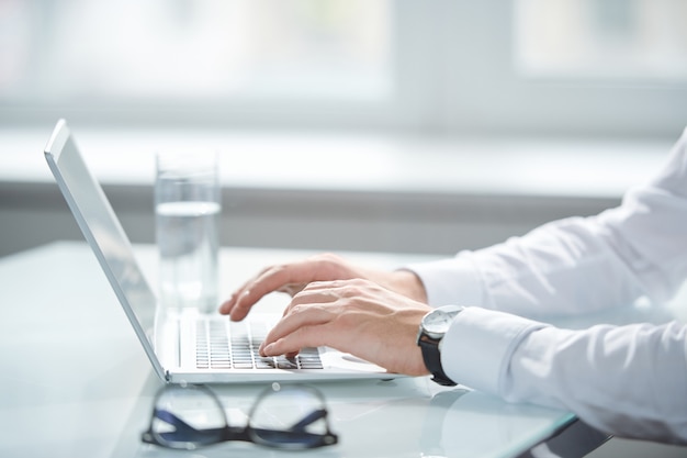 Mani di giovane uomo d'affari in camicia bianca toccando i tasti della tastiera mentre si utilizza il computer portatile per organizzare il lavoro