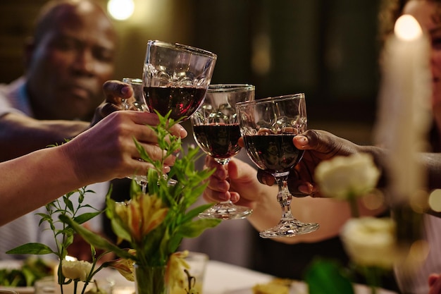 Mani di persone interculturali giovani e mature che tintinnano con bicchieri di vino pieni di vino rosso sul tavolo servito con piante verdi in vasi durante la cena