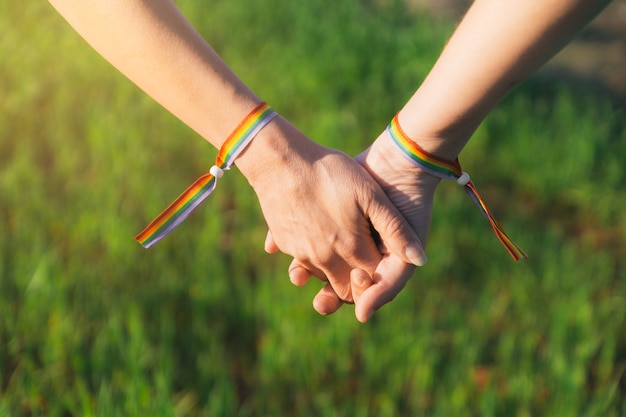 Mani di ragazze lesbiche giunte con braccialetti arcobaleno sul muro di erba verde sfocato alla retroilluminazione del tramonto