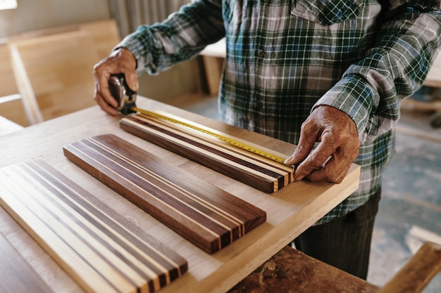 Mani di un anziano carpentiere che misurano pezzi di legno di noce e acero fusi insieme per realizzare un tagliere a grana tagliente