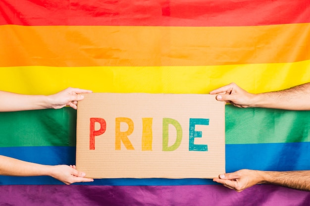 Mani di un uomo e di una donna che tiene un cartone con la parola orgoglio scritta a colori con la bandiera del gay pride dietro