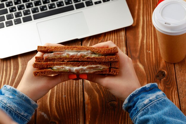 Mani di una donna che tiene il panino sopra il tavolo di lavoro con il computer portatile