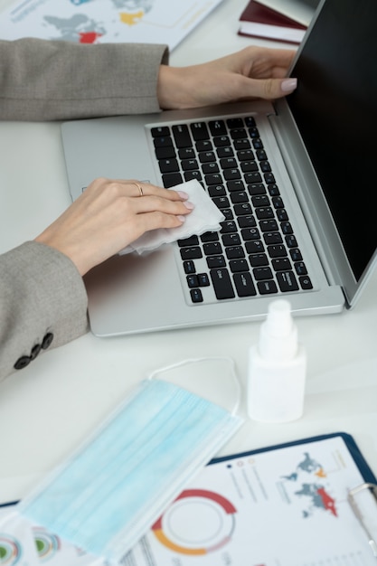 Mani di una giovane donna d'affari contemporanea in abiti da cerimonia grigi che puliscono la tastiera del laptop con disinfettante mentre sono seduti sul posto di lavoro in ufficio