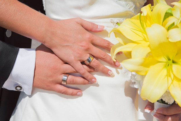 Mani di una sposa e sposo con fedi nuziali e bouquet di gigli gialli