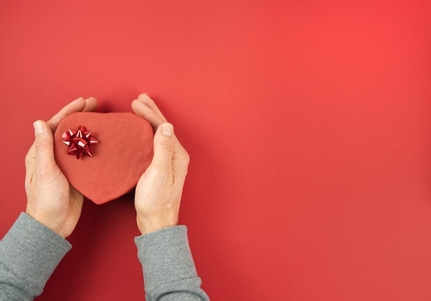 Mani di uomini che tengono confezione regalo a forma di cuore chiusa con ornamento su sfondo rosso. giorno di San Valentino