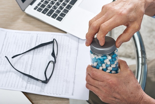 Mani di uomo anziano malato scoprendo vaso con pillole per prendere medicine mentre era seduto a tavola durante il lavoro con i documenti