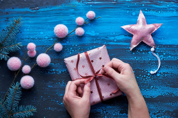 Mani femminili che avvolgono regalo di Natale, decorazioni invernali e ramoscelli di abete. Natale piatto giaceva in blu, nero e rosa su sfondo scuro liquido acrilico liquido vernice. Buon Natale e Felice Anno Nuovo 2020!