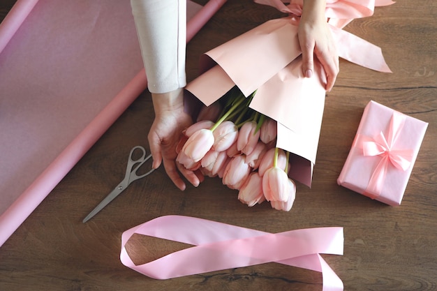 Mani femminili che avvolgono un mazzo di tulipani rosa in carta kraft rosa accanto a forbici e corda di iuta su tavolo di legno, vista dall'alto. Composizione piatta per creare bouquet di fiori freschi.