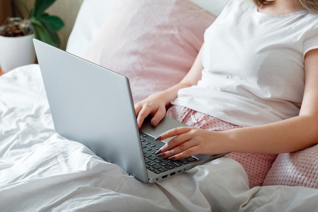 Mani femminili che digitano sulla tastiera del laptop mentre si trovano a letto. La donna lavora utilizzando il computer portatile mentre è seduta a letto a casa la mattina. Ragazza teenager in pigiama rosa che studia online o che pianifica la sua giornata.