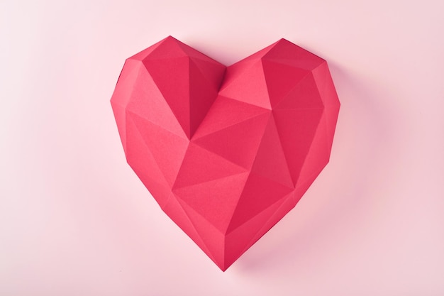 Mani femminili che tengono a forma di cuore di carta poligonale rossa su sfondo rosa. Cuore di carta poligonale rosso per San Valentino o altri biglietti d'invito d'amore. Vista dall'alto. Immagine tonica.