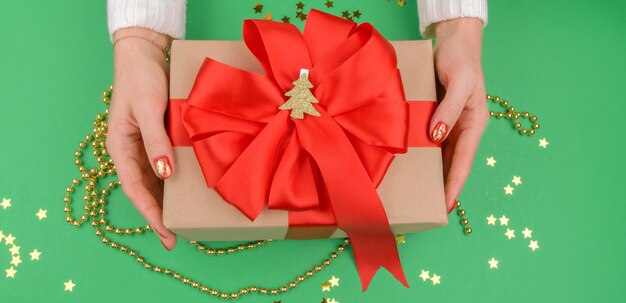 Mani femminili che tengono un regalo di Natale con fiocco rosso su sfondo verde con decorazioni dorate