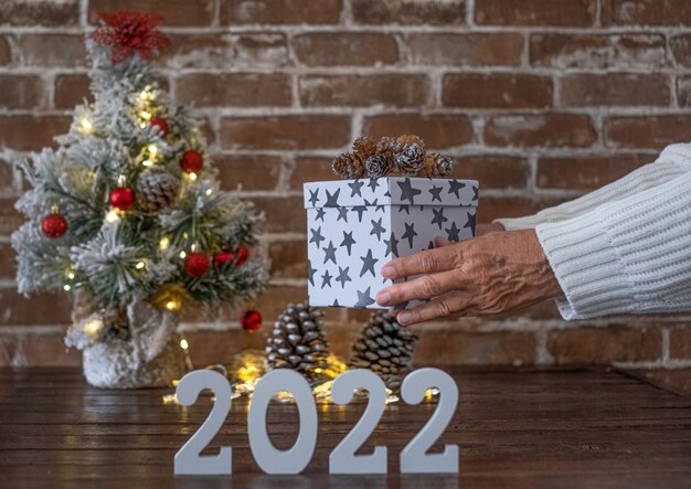 Mani femminili che tengono un regalo di Natale sul numero 2022. Buon Natale e felice anno nuovo, piccolo albero di Natale sul tavolo di legno con palline rosse e luci.