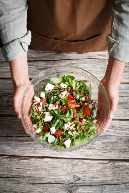 Mani femminili che tengono una ciotola di vetro di insalata di verdure con feta su uno sfondo di legno