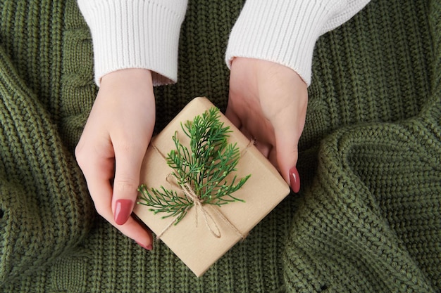 Mani femminili che tengono una confezione regalo kraft con decorazione di abete su maglione verde lavorato a maglia. Accogliente natura morta di Natale, compleanno o capodanno, regalo di San Valentino