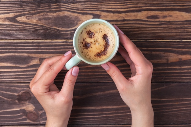 Mani femminili che tengono una tazza di caffè con schiuma sopra una tavola di legno, vista superiore. Latte o cappuccino con granelli di cioccolato.
