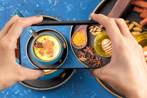 Mani femminili che tengono uno smartphone sopra una tazza di latte con latte di curcuma dorata con ingredienti in blu