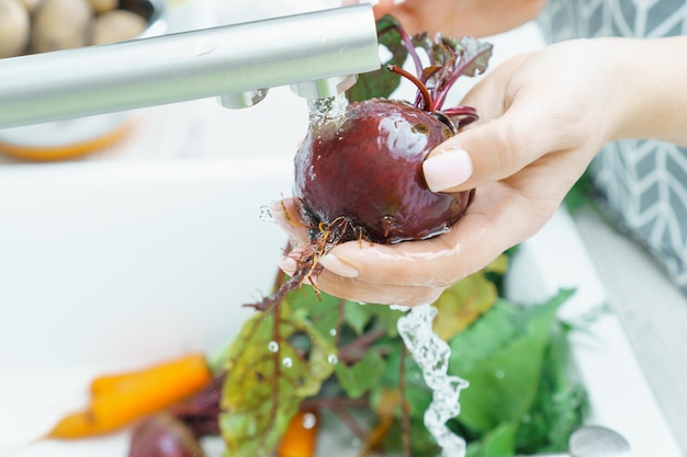 Mani femminili curate che lavano la barbabietola fresca sotto il getto d'acqua del rubinetto nel primo piano del lavello della cucina Barbabietole sporche con paglia verde