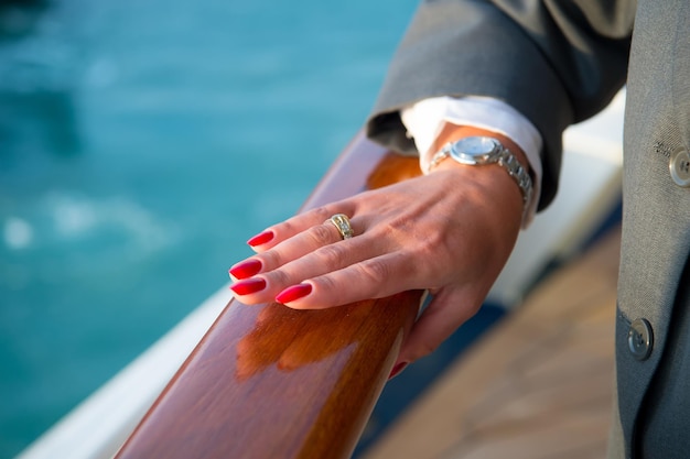 Mani femminili e orologio da polso con manicure rossa e anello dorato al dito con diamanti vicino all'acqua