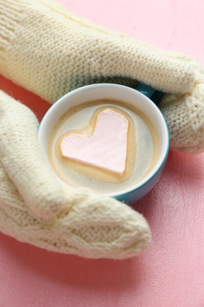 Mani femminili in guanti caldi che tengono una tazza di cappuccino caldo con cuore marshmallow su sfondo rosa da vicino