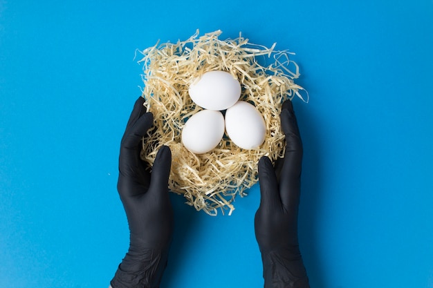 Mani femminili in guanti neri con uova di gallina bianche in un nido di paglia su sfondo blu