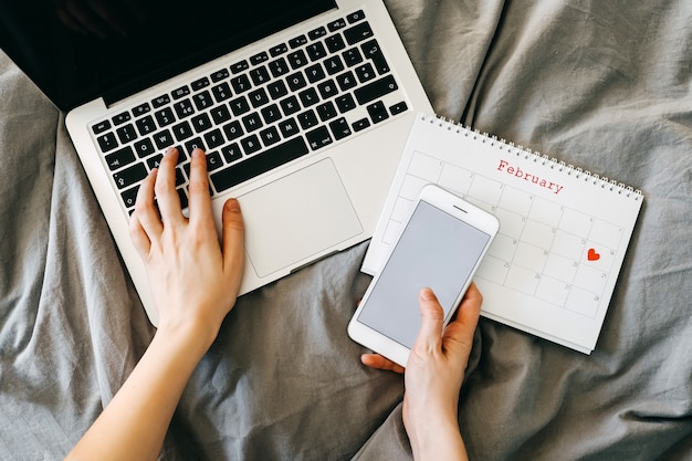 Mani femminili sul computer portatile, che tiene uno smartphone, con un calendario il giorno di San Valentino.