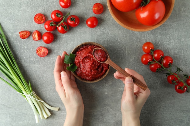 Mani femminili tengono ciotola e cucchiaio con concentrato di pomodoro su grigio con ingredienti