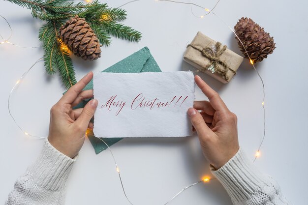 Mani femminili, vestite con un maglione bianco, che tengono in mano un foglio di carta bianca con la scritta Buon Natale.