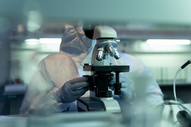 Mani guantate di scienziato in tuta usando il microscopio