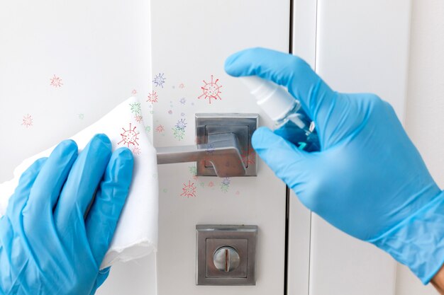 Mani in guanti protettivi - uno tiene lo spruzzatore con un antisettico, l'altro - pulisce la maniglia della porta con un panno inumidito con una soluzione disinfettante