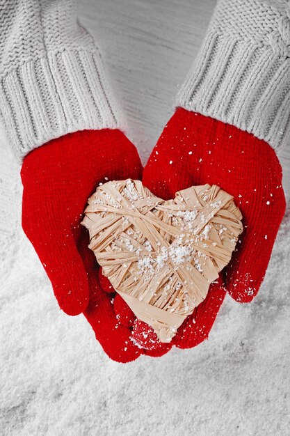 Mani in guanti rossi caldi che tengono il cuore di vimini su sfondo innevato