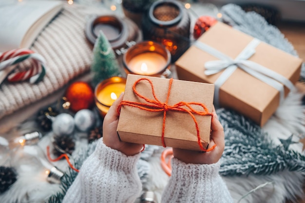 Mani in un maglione accogliente che apre il regalo di Natale con fiocco rosso sullo sfondo dell'albero di Natale con luci