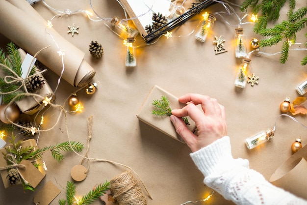 Mani in un maglione bianco confezionano un regalo per Natale e Capodanno in materiali ecologici: carta kraft, rami di abete vivo, pigne, spago. Etichette con mock up, decoro naturale, fatte a mano, fai da te. Atmosfera festosa