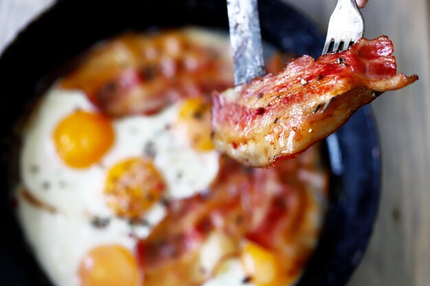 Mani maschii con un coltello e una forchetta stanno tagliando pancetta e uova in una padella.